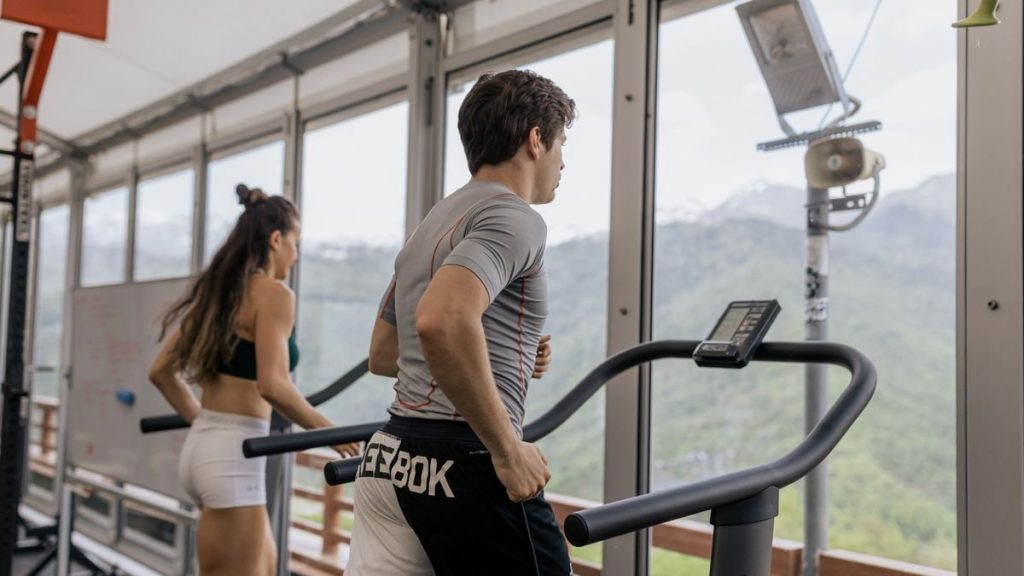 Two people running on treadmills in a gym with a mountain view, promoting fitness.