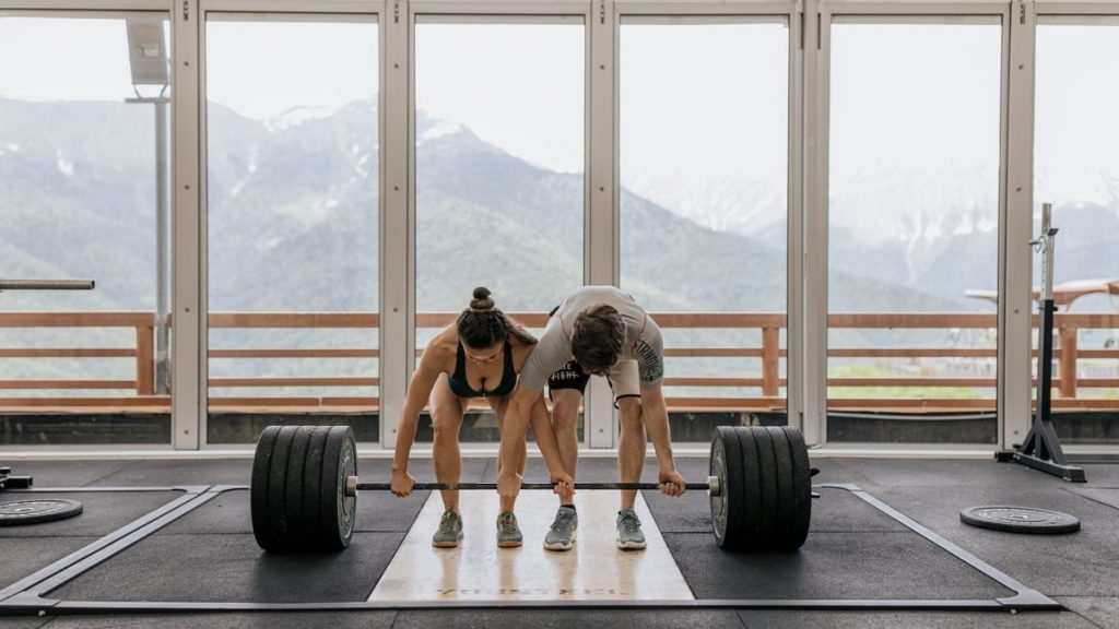A couple lifting a heavy barbell together in a gym with mountain views.