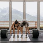 A couple lifting a heavy barbell together in a gym with mountain views.